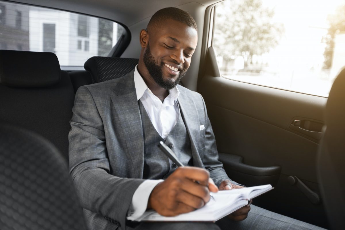 Happy black businessman sitting in car, taking notes